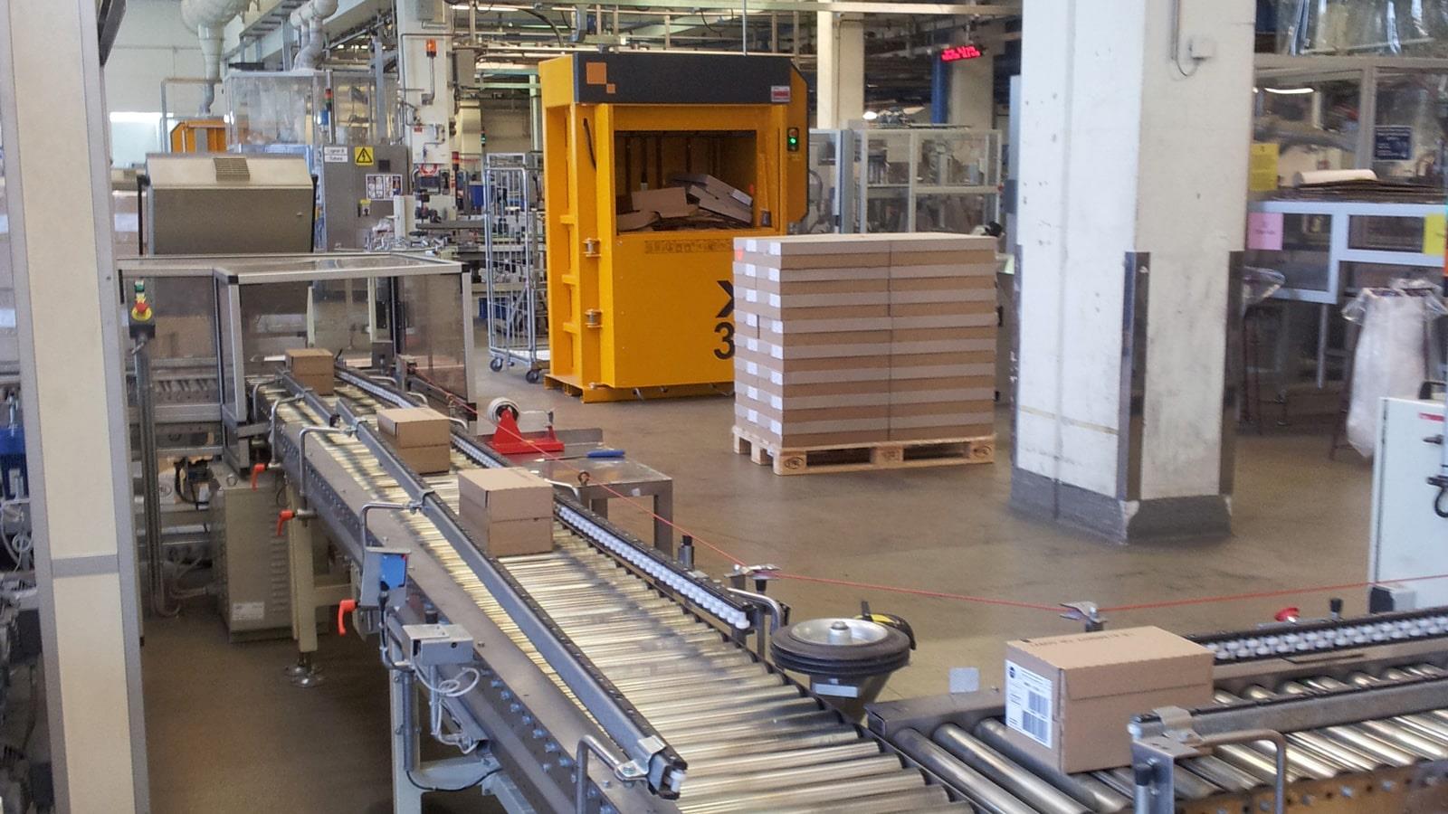 Roller conveyer with cosmetic in boxes next to baler with automatic door